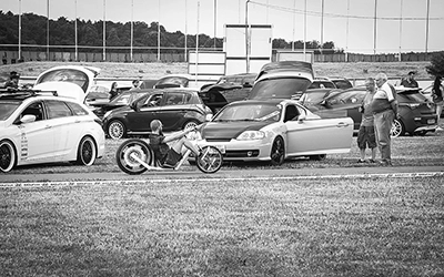 Ein Lowrider-Fahrrad mit Fahrer, im Vordergrund auf dem Gras, umgeben von verschiedenen Hyundai-Automodellen auf einem Treffen, in Schwarz-Weiß dargestellt. Fotograf Michael Schalansky.